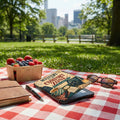 Picnic spread with fruit, e-reader, book, pen, and sunglasses on a checkered blanket.