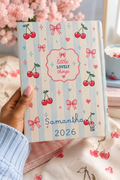 Notebook with cherry and bow design held by a hand, with 'little lovely things' text and 'Samantha 2026' on cover.