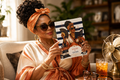 Hot Flashes and Plot Twists Menopause Book Club personalized hardcover journal held by mature African American woman in coral head wrap and tortoiseshell sunglasses in cozy living room with brass vintage fan